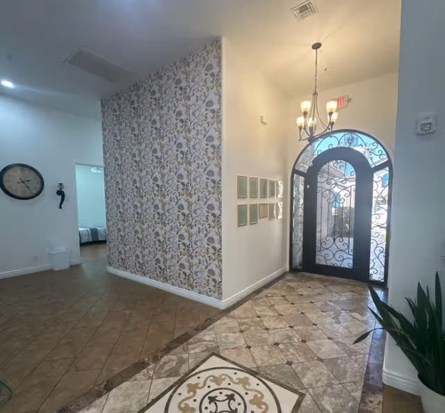 Bright entry foyer with patterned wallpaper, decorative tiled floor, an arched wrought-iron glass front door and a hanging chandelier.