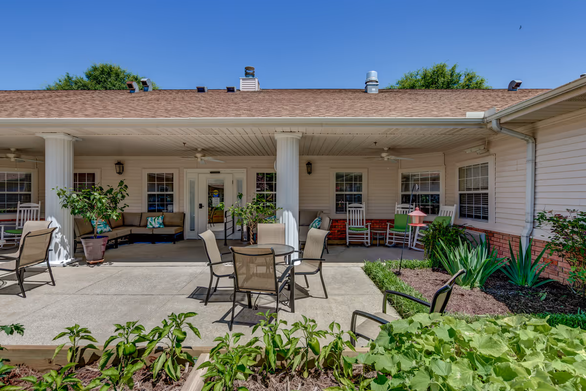 Outdoor patio area at Garrett Woods Senior Living Community with several chairs and tables arranged on a concrete surface. The patio is partially covered by a roof supported by white columns. There are potted plants and garden beds with green foliage in the foreground. The building exterior features beige siding with brick accents and multiple windows.