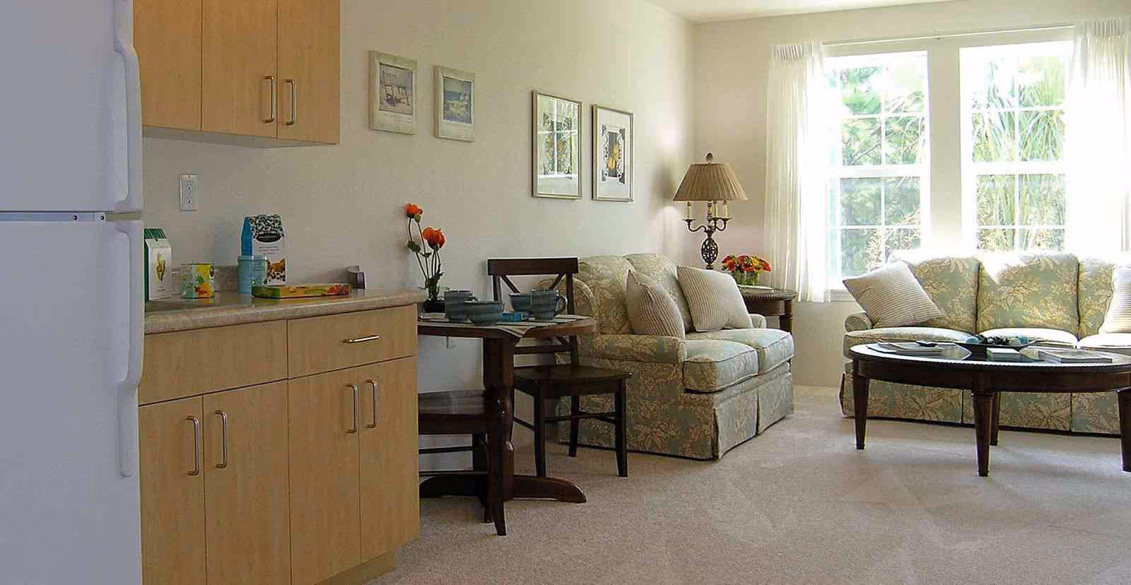 Sunlit living area with a small kitchenette on the left, floral sofas, a round coffee table, and a dining nook by a large window.