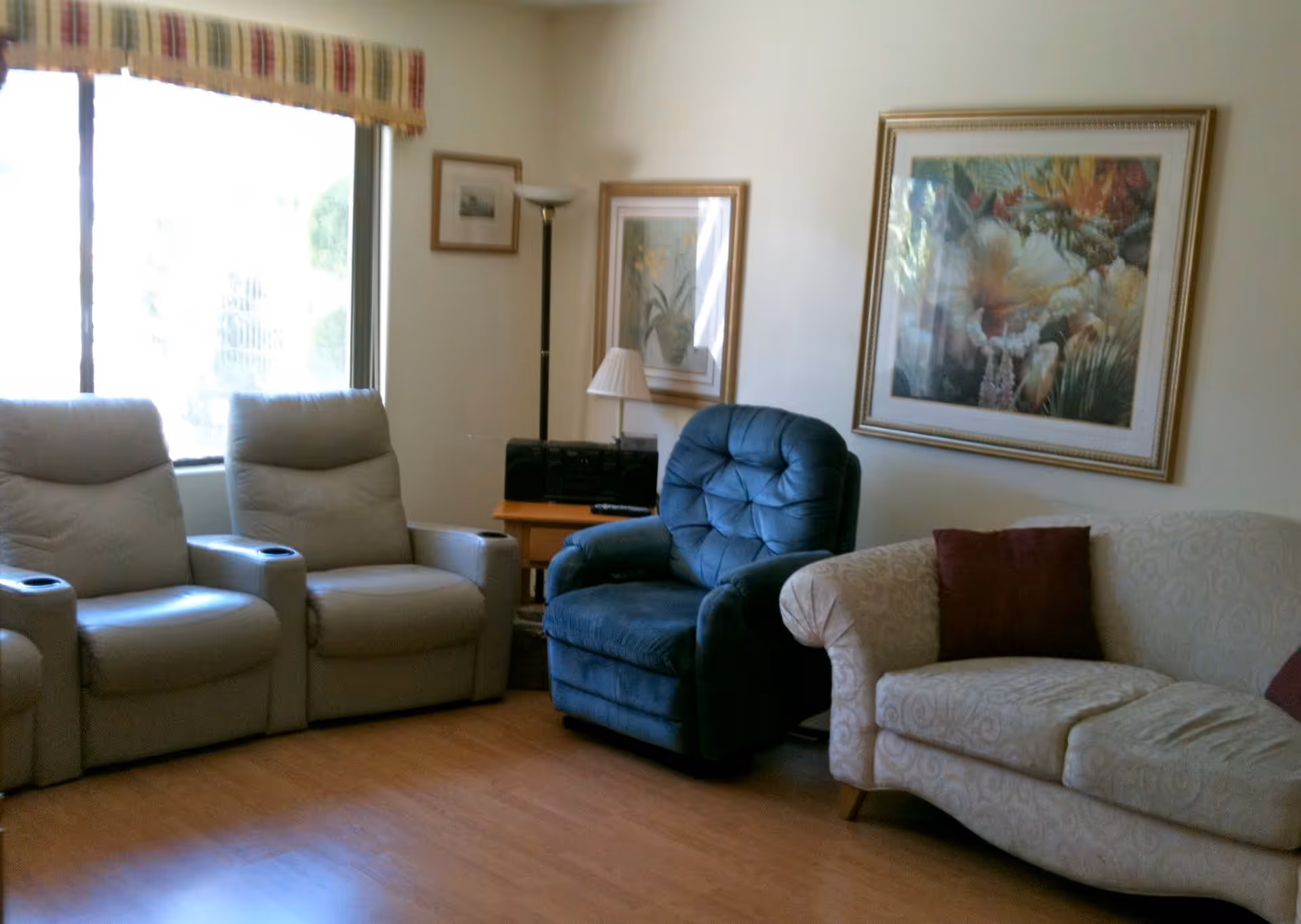 A cozy living room with two beige recliner chairs, a blue cushioned armchair, and a cream-colored sofa with a red pillow. The room has wooden flooring, a window with a striped valance, framed artwork on the walls, and a small wooden side table with a lamp and a stereo system.