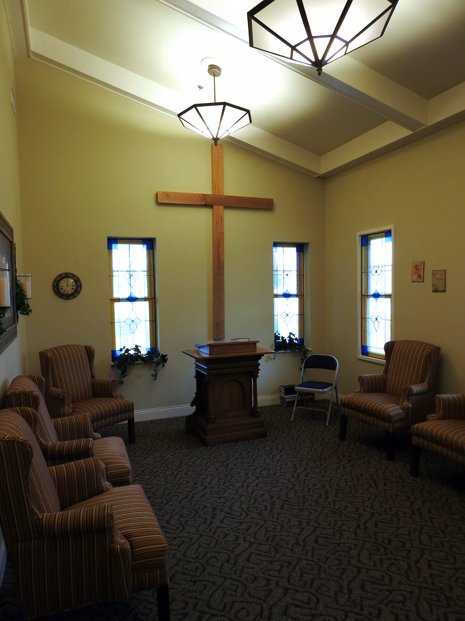 Interior room with a large wooden cross mounted on the wall between two stained glass windows. The room has a wooden podium in the center and several striped upholstered armchairs arranged around the perimeter. There is also a folding chair near the podium and two ceiling light fixtures.