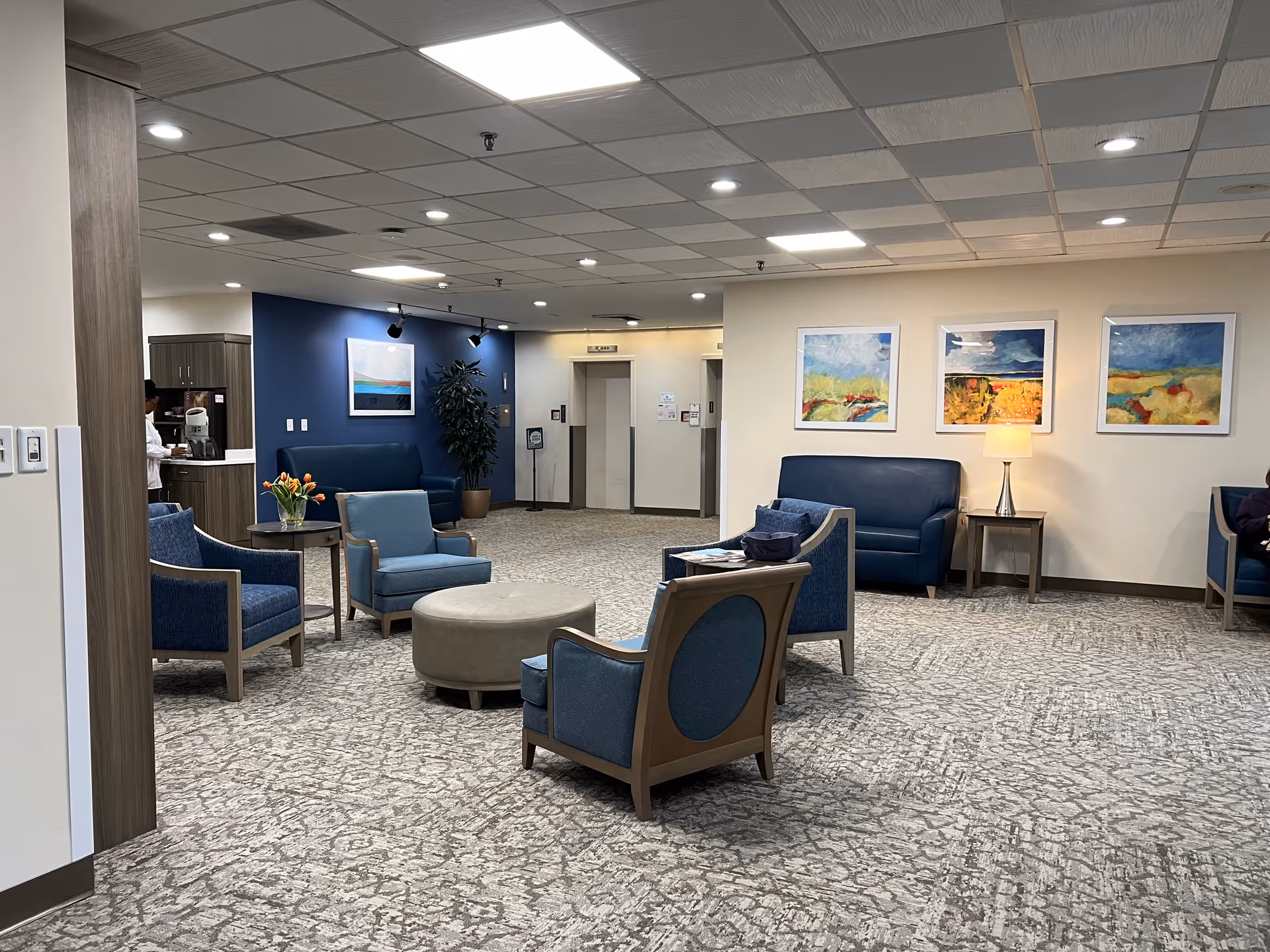 A spacious and well-lit common area in Myers Apartments Independent Living featuring multiple blue upholstered chairs and sofas arranged around a round ottoman. The room has a patterned carpet, a small table with a lamp, and colorful abstract paintings on the walls. In the background, there are two elevators and a small kitchenette area with a person preparing coffee.