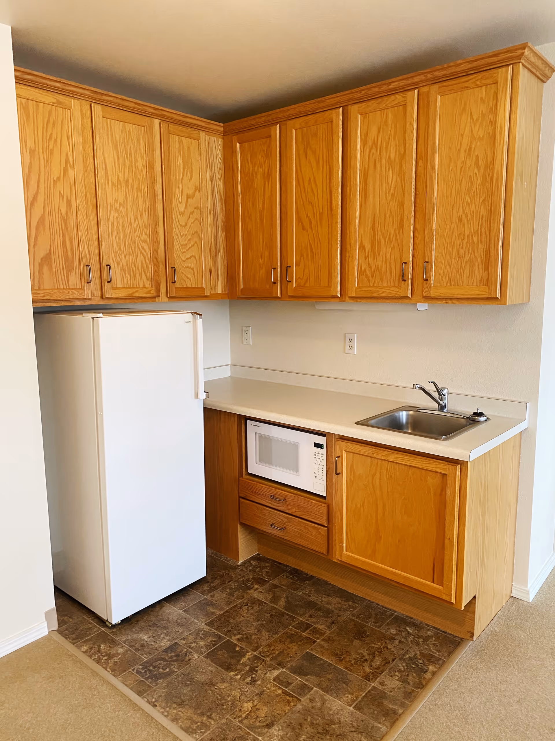 Small kitchen area with wooden cabinets, a white refrigerator, a white microwave built into the lower cabinets, a countertop with a stainless steel sink and faucet, and a tiled floor section in front of the kitchen.