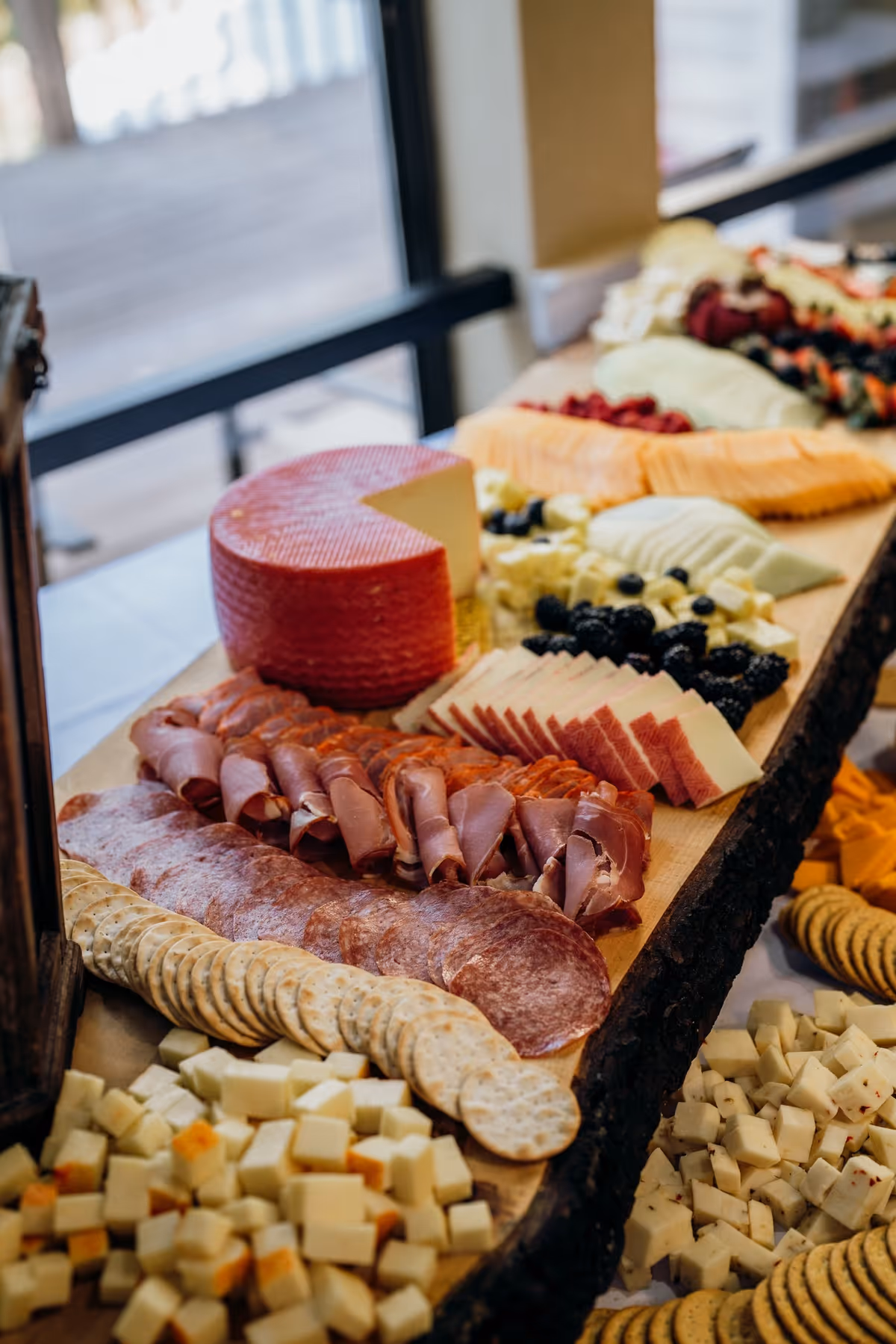 A charcuterie spread of sliced meats, cheeses, crackers, and fruit arranged on a long wooden board near a window.