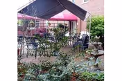 Outdoor courtyard with tables and chairs under a canopy and pink umbrella, surrounded by plants and a brick building.