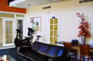 Interior view of a senior living facility lounge area with a black leather couch, exercise bike, two wooden chairs, a glass table with a telephone, and a tall vase with red and orange artificial flowers against a beige wall with two white doors and a bulletin board.