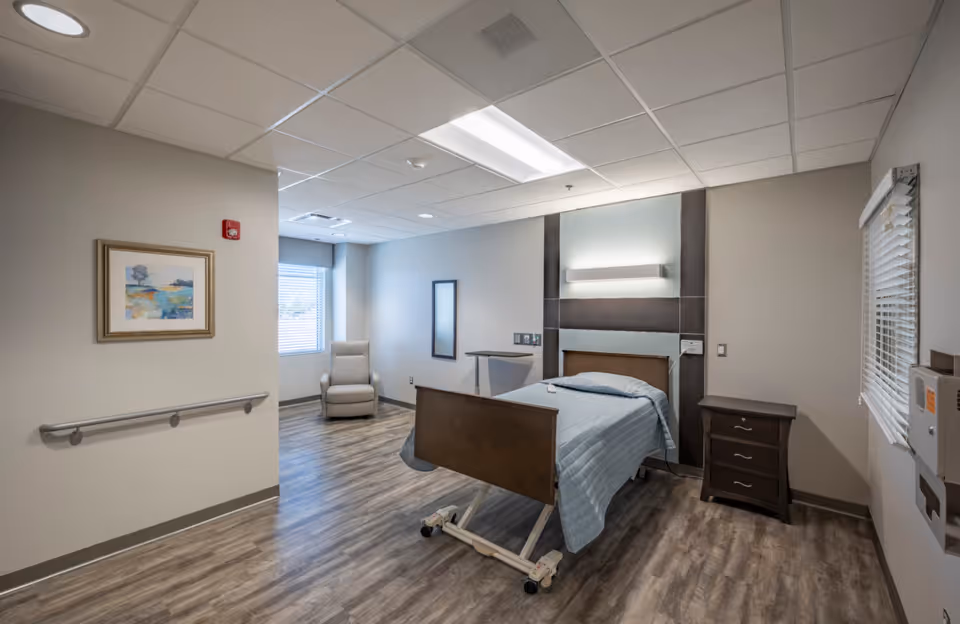 A clean and well-lit senior living bedroom with a single hospital-style bed covered with a light blue blanket, a wooden nightstand with three drawers, a beige recliner chair near a window with blinds, a framed abstract painting on the wall, and a wall-mounted light fixture above the bed. The room has wood-look flooring and neutral-colored walls.