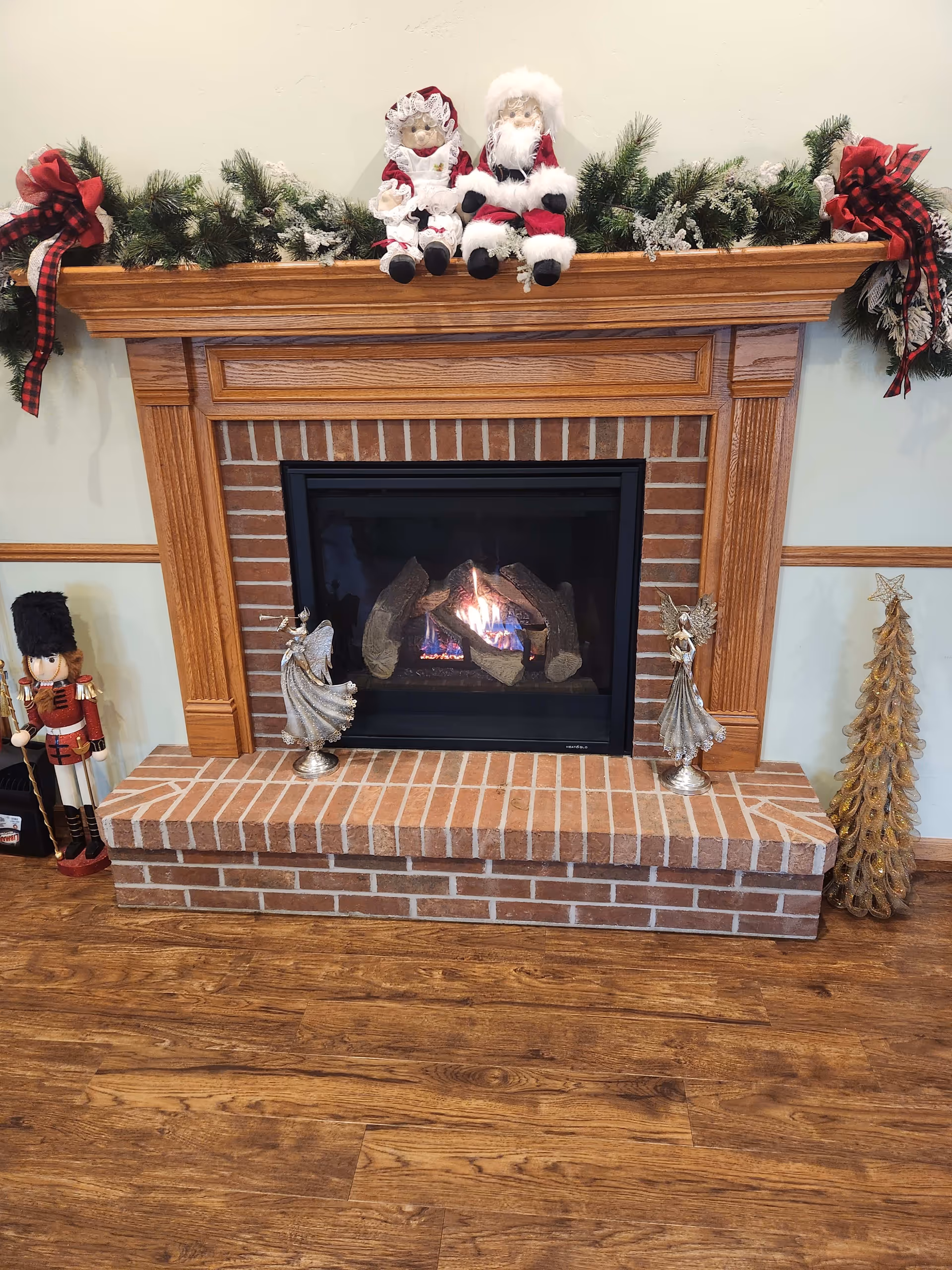 A cozy brick and wood fireplace decorated for Christmas with a garland and red plaid bows on the mantle. Two festive dolls dressed as Santa Claus and Mrs. Claus sit on the mantle. On the hearth, there are two silver angel figurines, a nutcracker figure on the left, and a gold Christmas tree decoration on the right. The floor is wooden.