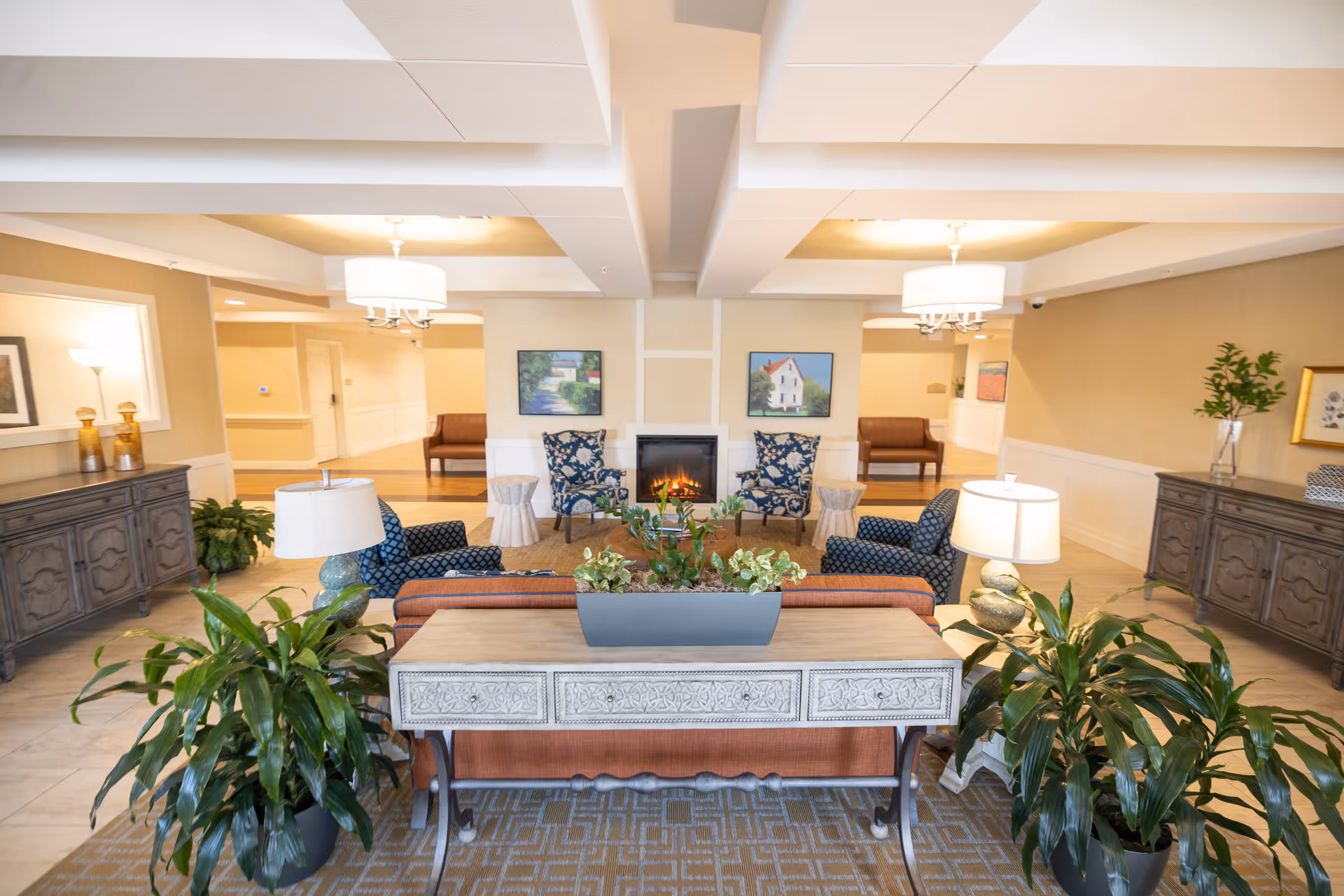 A bright and spacious senior living facility common area with a cozy fireplace in the center. The room features comfortable seating including patterned armchairs and a sofa, two side tables with lamps, decorative plants, and framed artwork on the walls. The ceiling has recessed lighting and chandeliers, and there are two wooden sideboards along the walls.