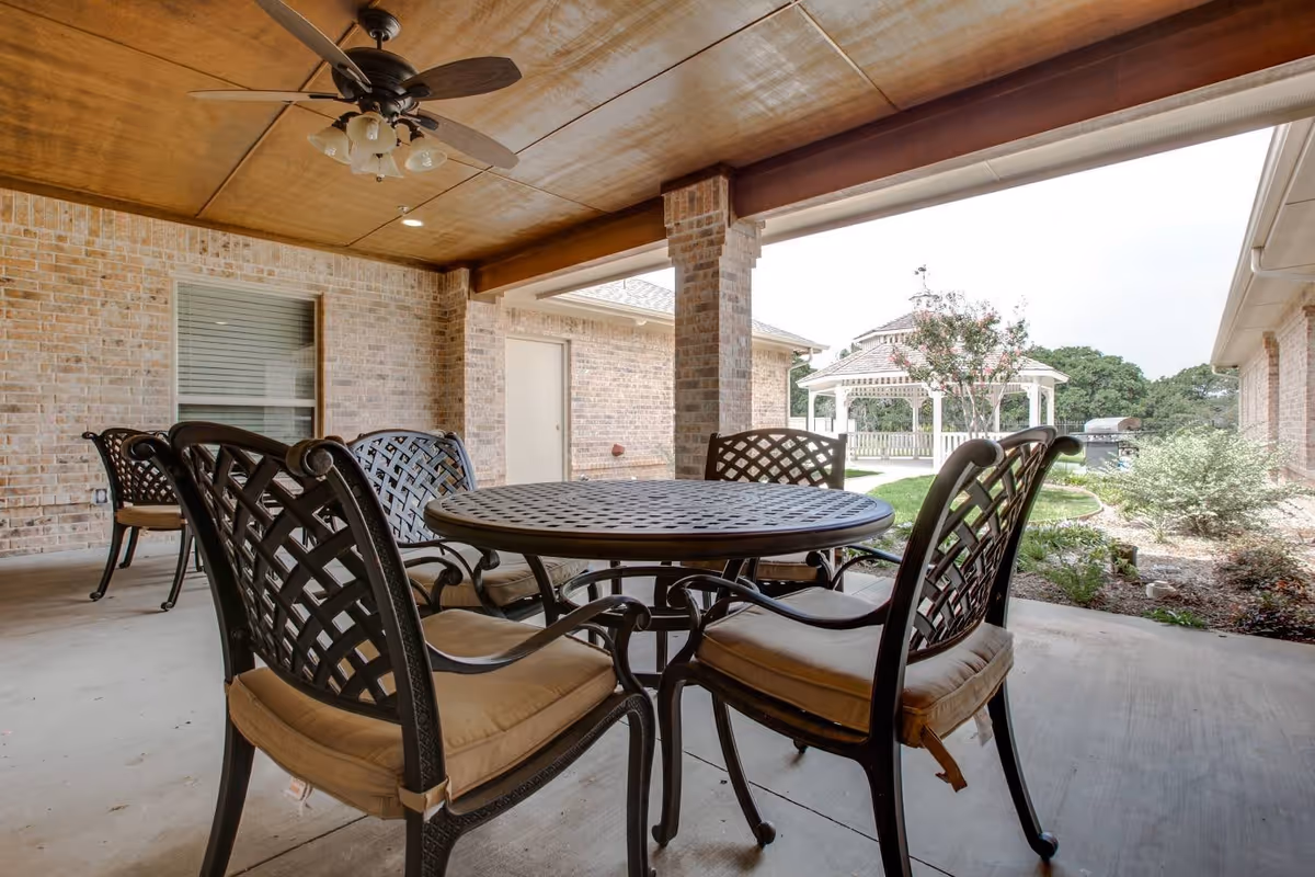 Covered outdoor patio area with a round metal table and six cushioned chairs. The ceiling is wooden with a ceiling fan and light fixture. In the background, there is a brick wall with a window and door, and beyond the patio, a garden area with a white gazebo and greenery is visible.