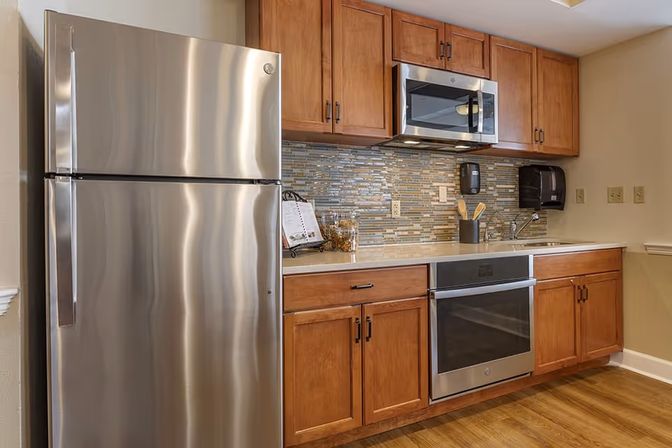 A modern kitchen area featuring stainless steel appliances including a refrigerator, microwave, and oven. The kitchen has wooden cabinets, a tiled backsplash in shades of gray and beige, a countertop with kitchen utensils, and a sink. The floor is wooden.