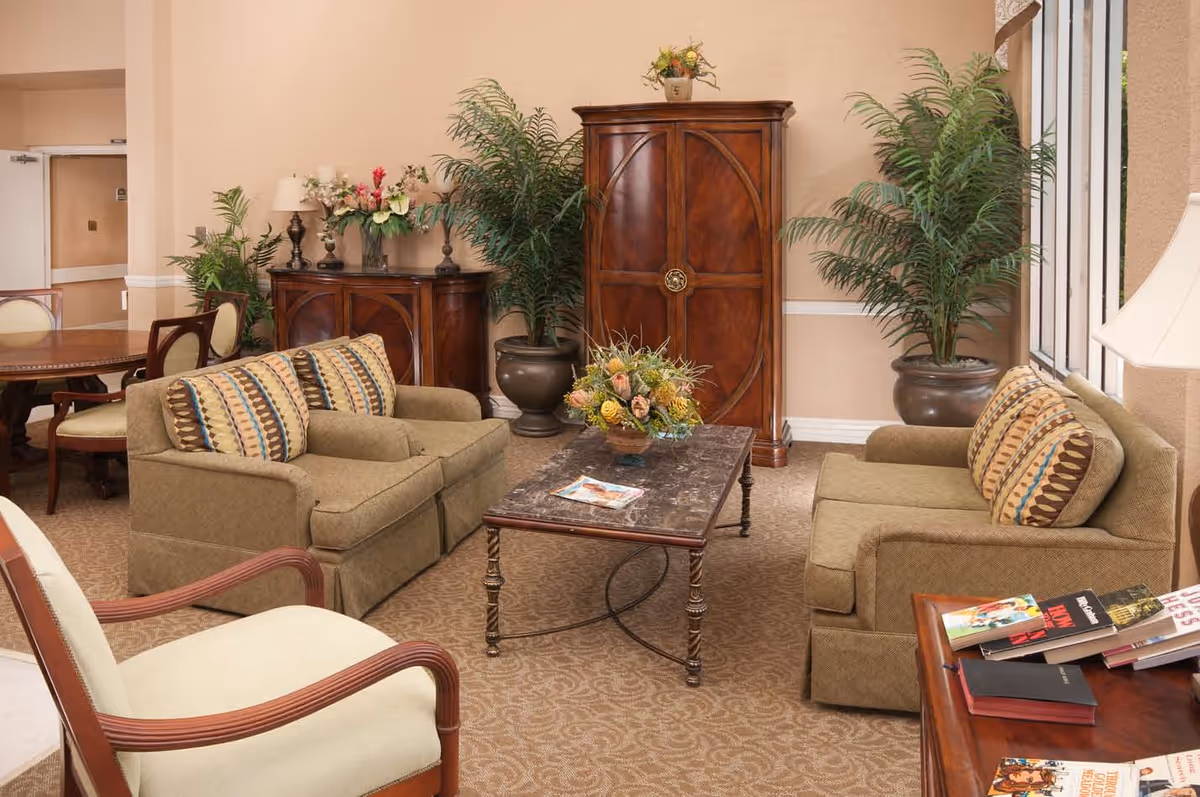 A cozy living room area with two beige sofas adorned with patterned cushions, a marble-top coffee table with a floral centerpiece, a wooden cabinet, potted plants, and a side table with books. The room has beige walls and carpet, with a large window letting in natural light.