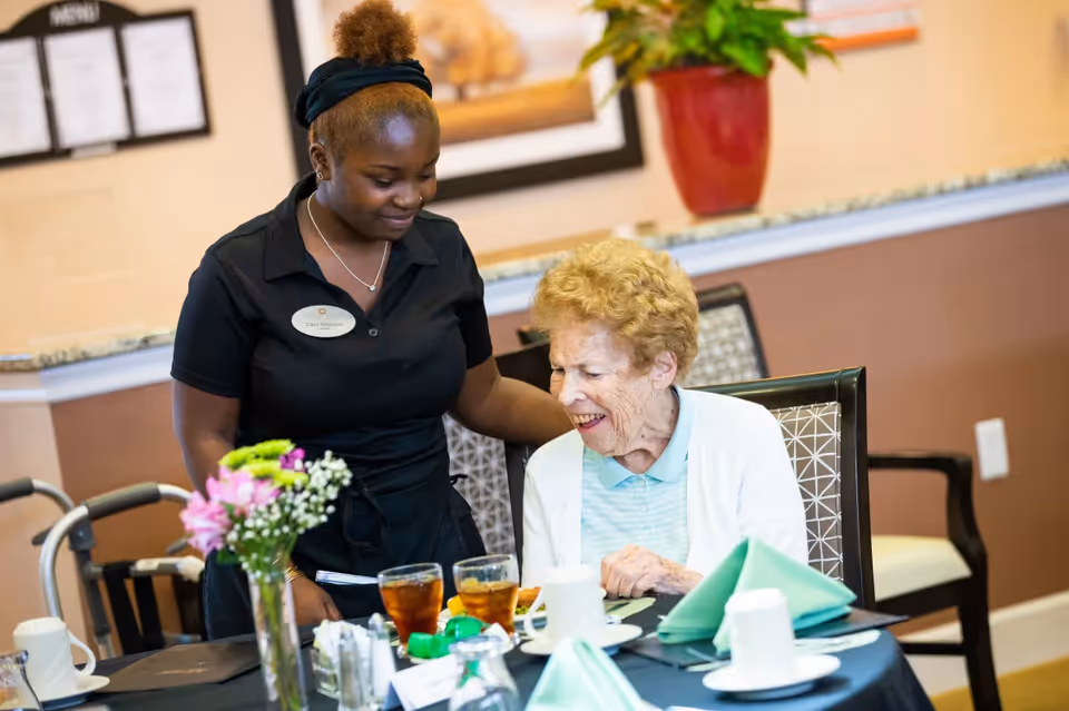 A smiling elderly woman seated at a dining table with two glasses of iced tea and green folded napkins, being assisted by a smiling staff member wearing a black uniform and name tag in a warmly decorated dining area.