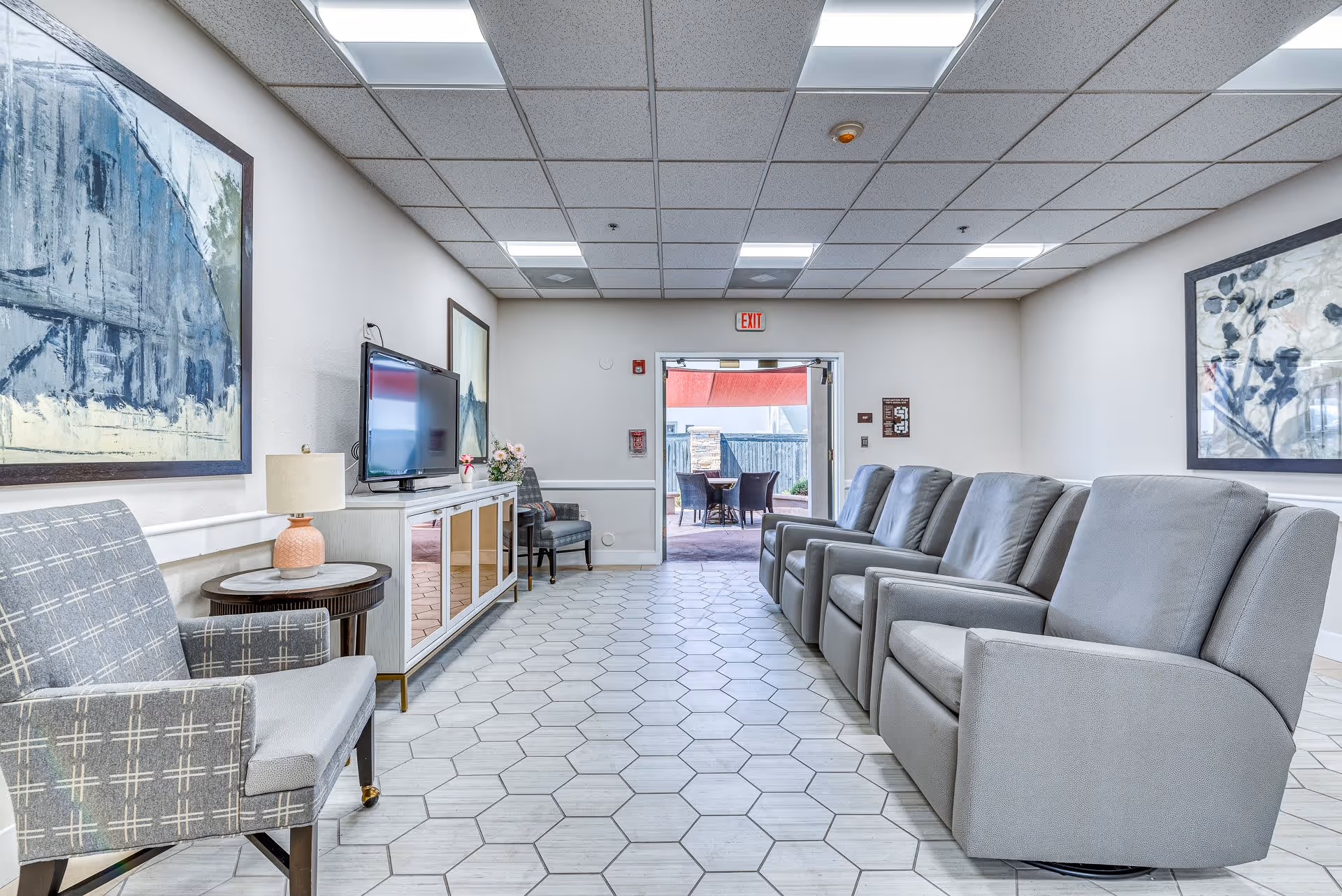 A bright and clean lounge area with gray cushioned armchairs lined up on the right side and a patterned armchair on the left. A flat-screen TV is mounted on a white cabinet against the left wall, with a small table and lamp beside it. Large abstract paintings hang on the walls, and the room has hexagonal tile flooring. At the far end, a door opens to an outdoor patio with tables and chairs under a red umbrella.