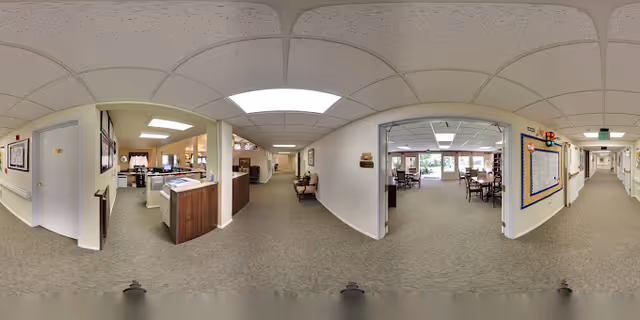 Panoramic view of an interior senior living facility hallway with carpeted floors and white walls. On the left side, there is a reception desk area with office equipment and chairs. Further down the hallway, there are tables and chairs arranged in a common area with large windows letting in natural light. The right side shows a bulletin board and doors leading to other rooms. The ceiling has recessed lighting and acoustic tiles.