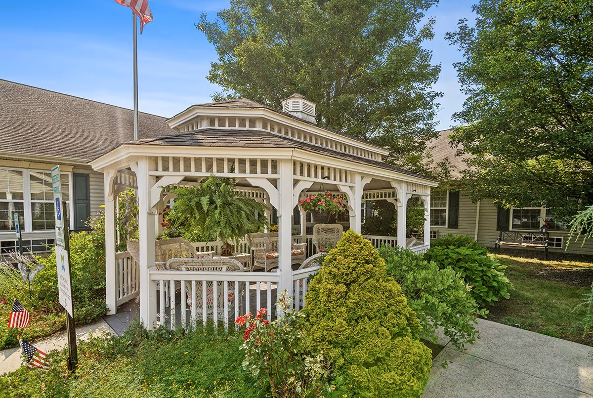 A white wooden gazebo with wicker chairs and hanging plants, surrounded by green bushes and trees, located outside a building with beige siding and green shutters under a clear blue sky.