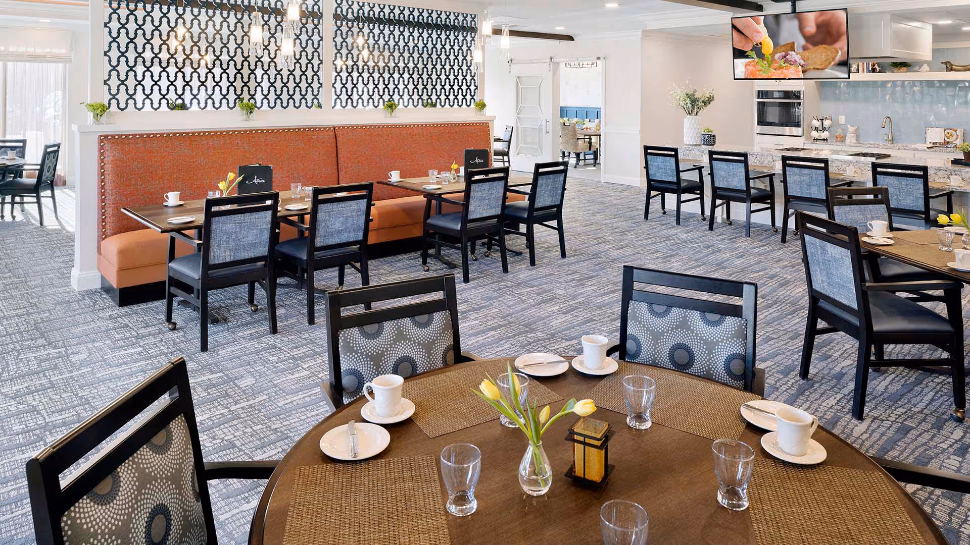 A bright and spacious dining room with multiple tables and chairs arranged neatly. The tables are set with cups, plates, glasses, and small vases with yellow tulips. There is a long orange cushioned bench along one wall with black chairs and tables in front of it. The room features a patterned carpet, decorative wall panels, and a kitchen area with a countertop and stools in the background. A TV mounted on the wall shows a close-up of food preparation.