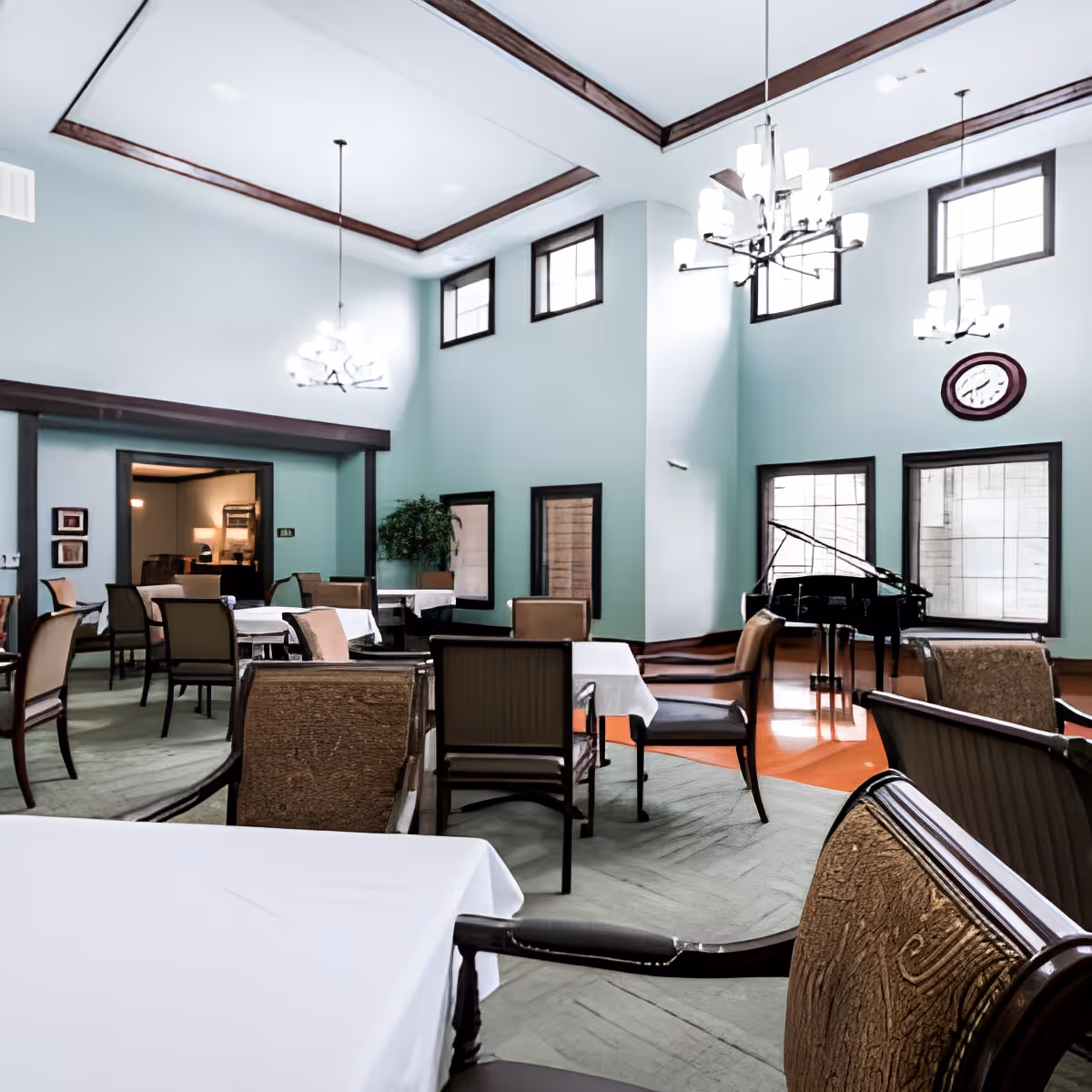A spacious dining room with multiple tables covered in white tablecloths and surrounded by chairs. The room features high ceilings with wooden trim, large windows, modern chandeliers, a clock on the wall, and a grand piano in the corner.