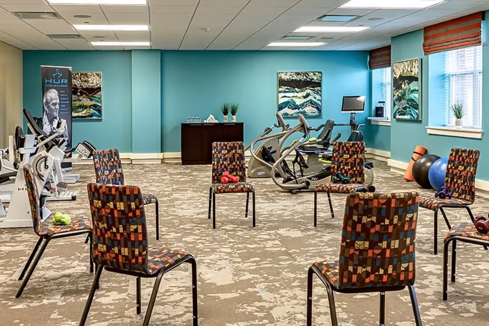 A fitness room with exercise equipment including stationary bikes and elliptical machines. Several colorful patterned chairs are arranged in a circle in the center of the room, each with small dumbbells placed on or near them. The walls are painted teal and decorated with abstract artwork. Large windows with red blinds allow natural light into the space.