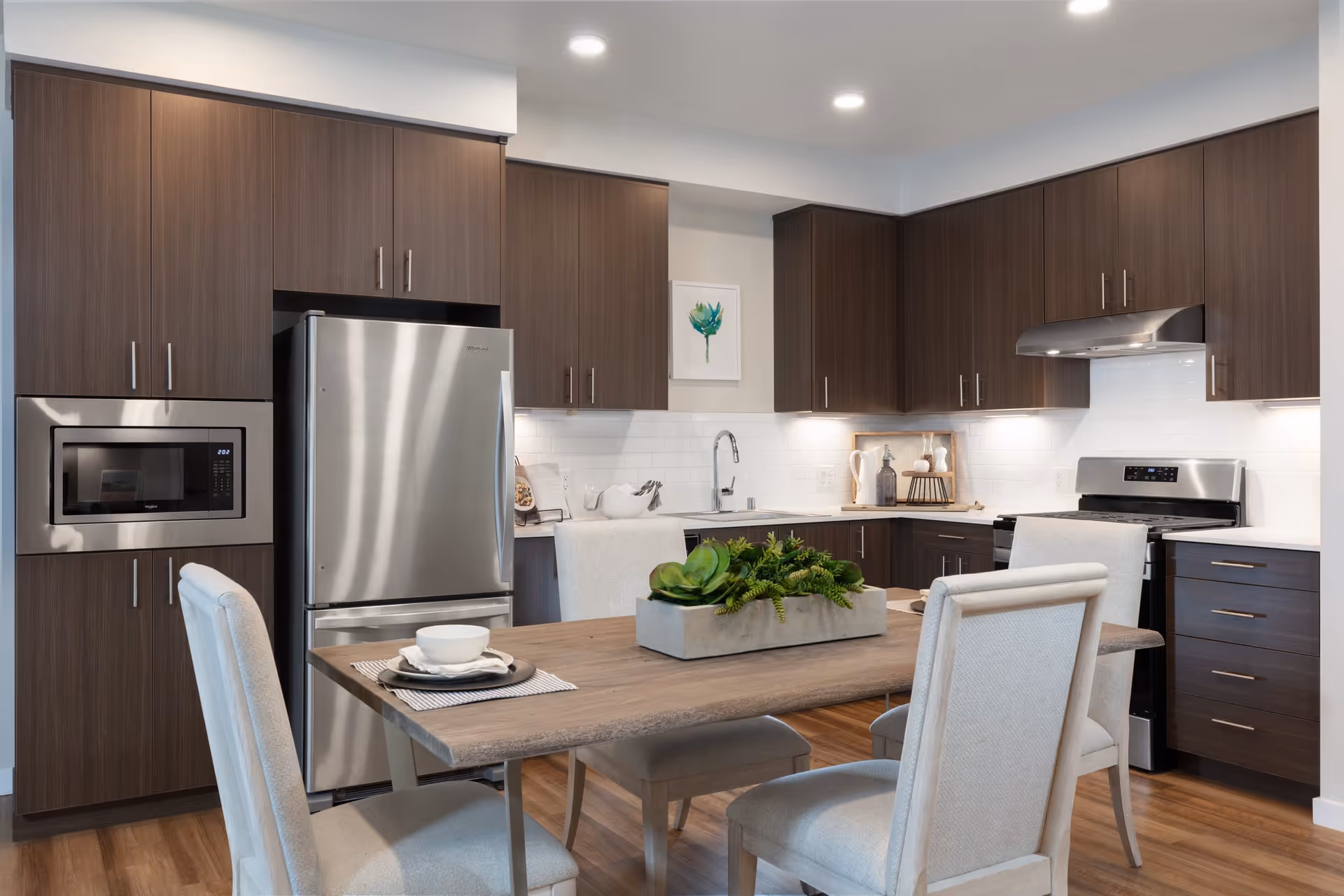 Modern kitchen with dark wood cabinets, stainless steel refrigerator, microwave, and stove. A wooden dining table with four upholstered chairs is in the foreground, decorated with a rectangular planter of green succulents. The kitchen has white tiled backsplash and under-cabinet lighting.