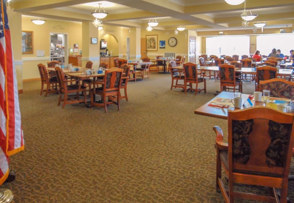 A spacious dining room with multiple wooden tables and chairs arranged neatly. The room has beige walls, carpeted floors, and ceiling lights with decorative snowflakes hanging. An American flag is visible on the left side, and a few people are seated at tables near large windows letting in natural light.