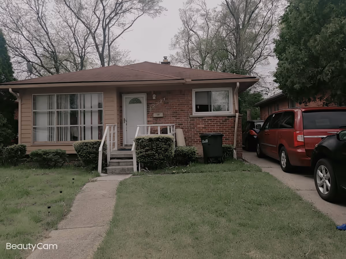 Front exterior view of a single-story brick and siding house with a small porch, steps leading up to the front door, bushes in front, and a driveway with parked cars on the right side. Trees are visible in the background under an overcast sky.