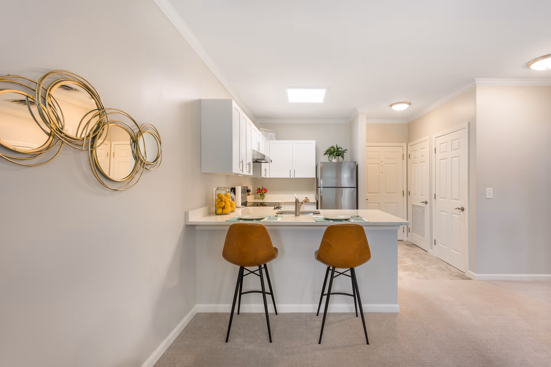 Modern kitchen area with white cabinets, a stainless steel refrigerator, and a countertop with two brown bar stools. The countertop has place settings and a glass container filled with lemons. A decorative circular mirror hangs on the wall to the left. The area is well-lit with ceiling lights and has beige walls and carpeted flooring.
