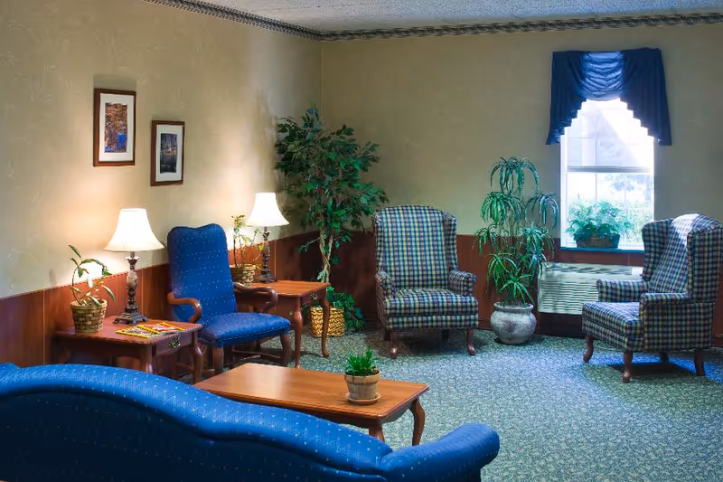 Sunlit common living room with upholstered armchairs, a sofa, coffee tables, lamps, and potted plants.