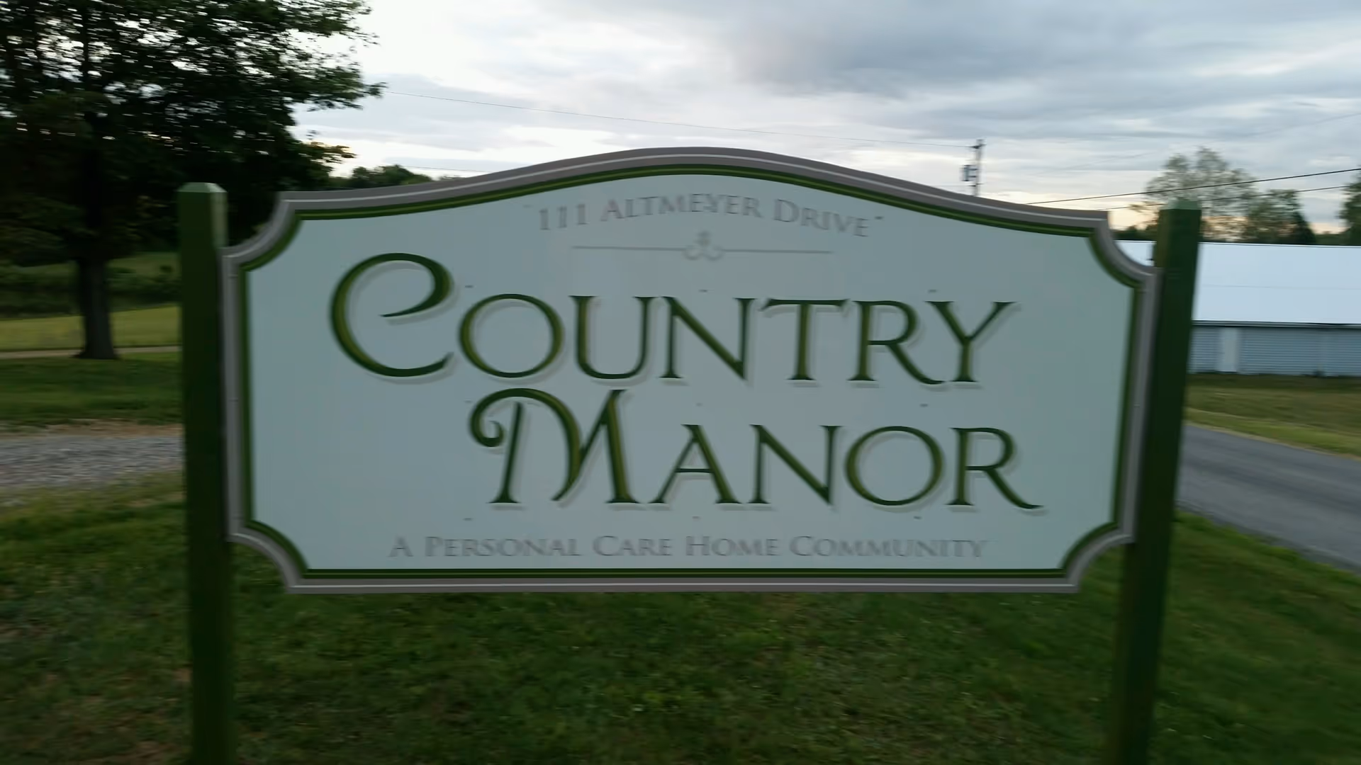 A green and white outdoor sign for Country Manor, a personal care home community, located at 111 Altmeyer Drive, with grass, trees, and a road in the background under a cloudy sky.