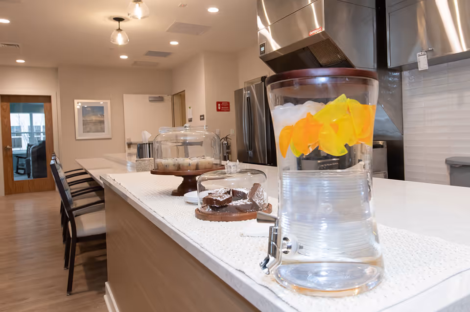 A modern kitchen area with a long white countertop featuring a large glass dispenser filled with water and lemon slices, a covered plate of brownies, and a covered plate of muffins. In the background, there are stainless steel appliances, including a refrigerator and a range hood, with wooden chairs lined up along the counter.