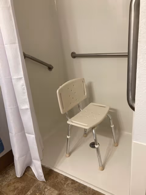 A shower area with a white plastic shower chair with metal legs, grab bars on the walls for support, and a white shower curtain partially visible on the left side.