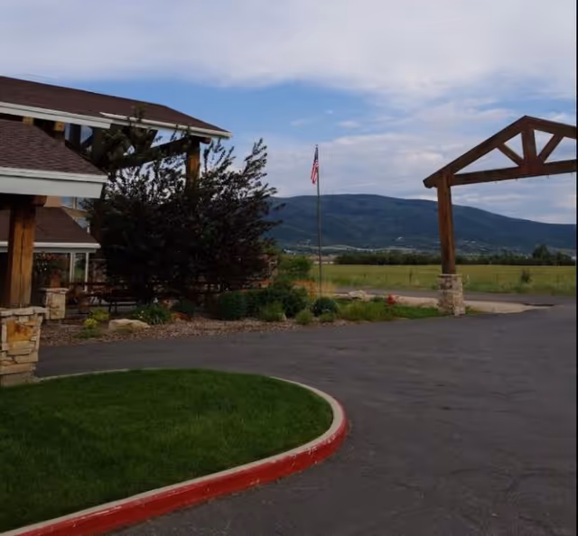 Driveway and entrance area of an assisted living facility with a flagpole, wooden entry arch, lawn, and mountains in the background.