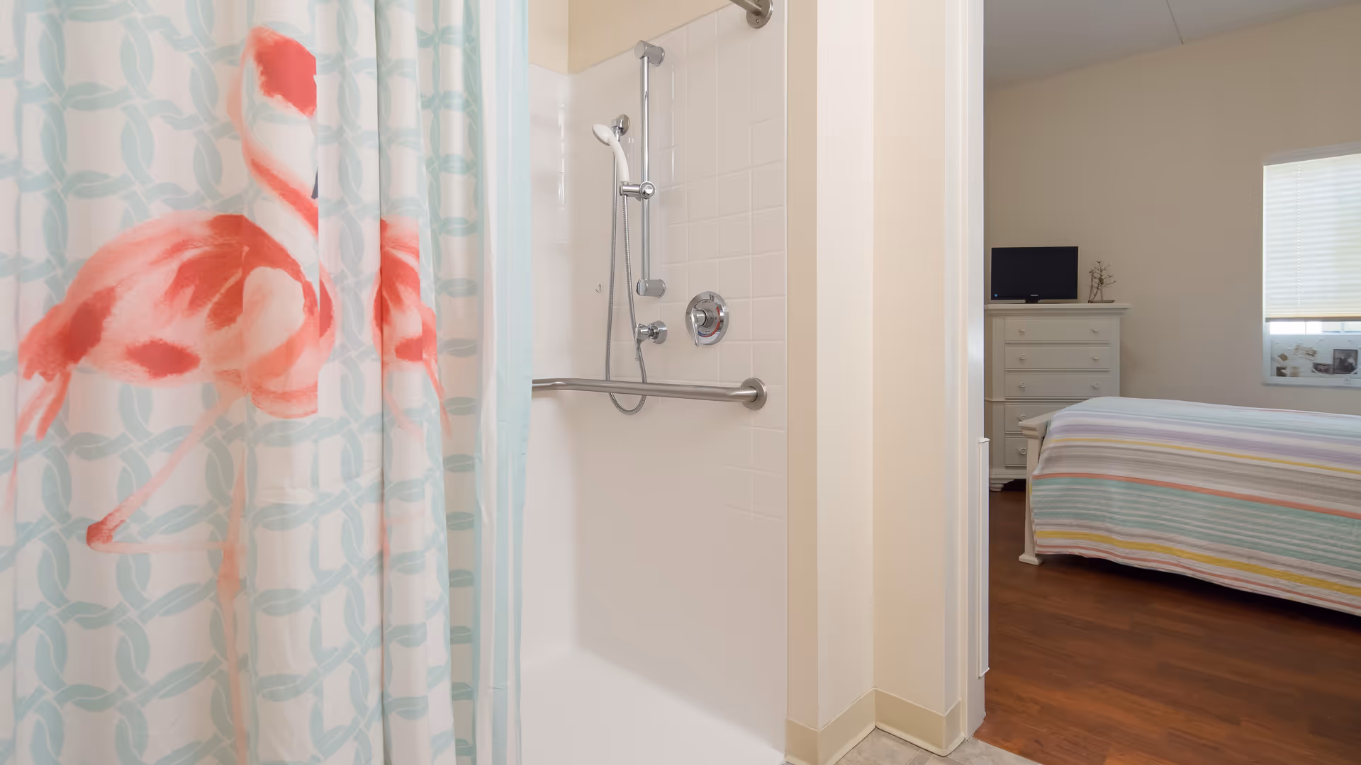 View of a bathroom shower with a white tiled wall and a silver grab bar, partially covered by a shower curtain featuring a pink flamingo design. Adjacent to the bathroom is a bedroom with a bed covered in a striped pastel bedspread, a white dresser with a small TV on top, and a window with blinds.