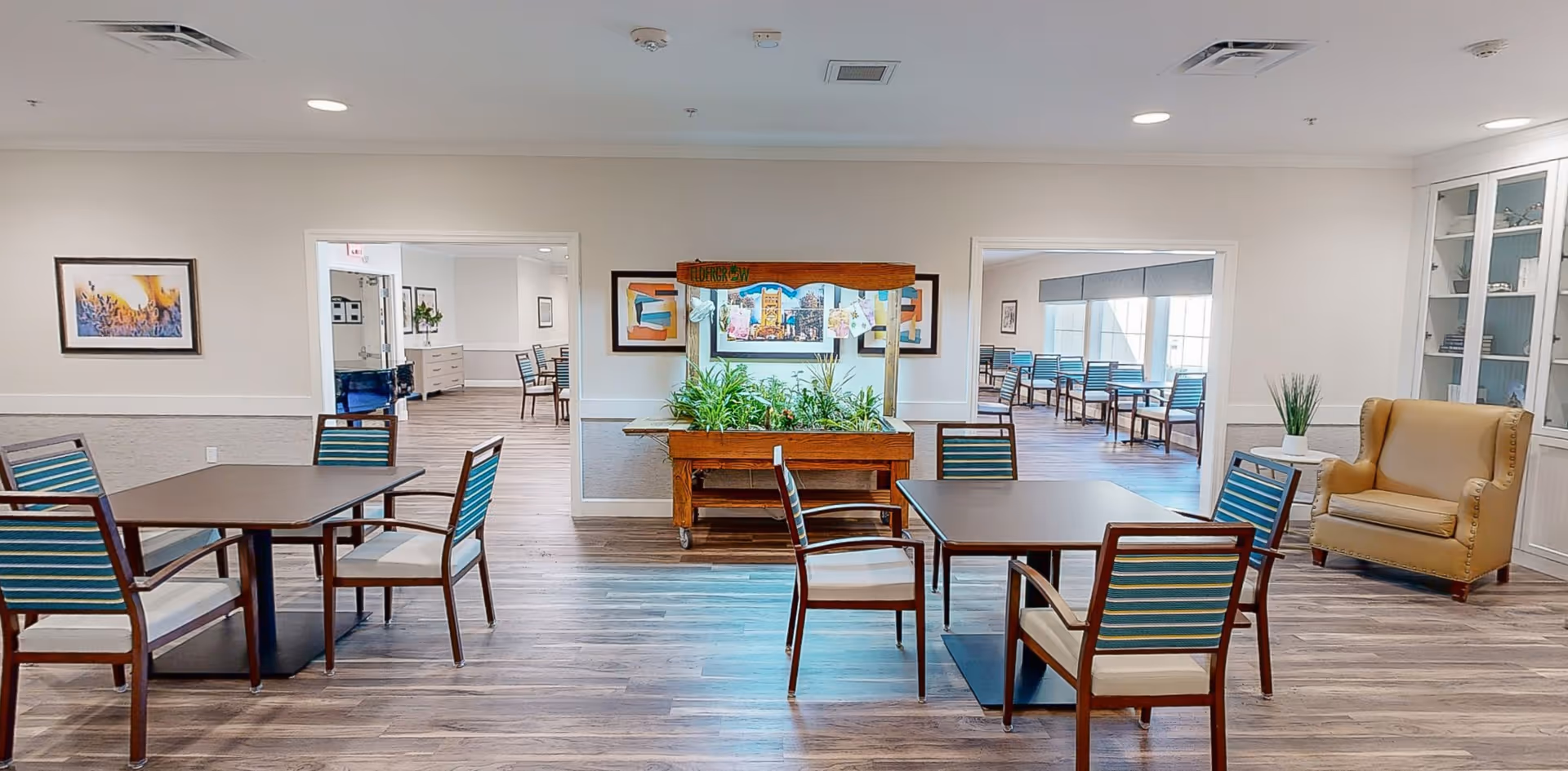 A bright and spacious common area in a senior living facility with wooden floors, several tables and chairs with striped cushions, a beige armchair, and a planter with green plants in the center. The room has white walls with framed artwork and two open doorways leading to additional seating areas with more tables and chairs.