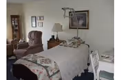 A cozy room with a single bed covered in a quilted bedspread, a bedside table with a lamp, a reclining chair, a small display cabinet, framed pictures on the wall, and a white desk with a chair.