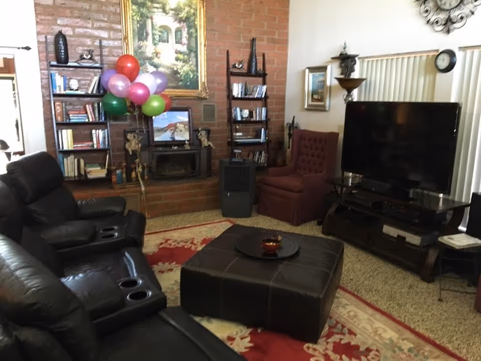 A cozy living room with black leather recliner sofas, a large ottoman in the center with a tray and bowl on top, a red patterned rug, a brick fireplace with a TV above it, bookshelves on either side filled with books, a cluster of colorful balloons, a maroon armchair, a large flat-screen TV on a wooden stand, and vertical blinds covering the window.