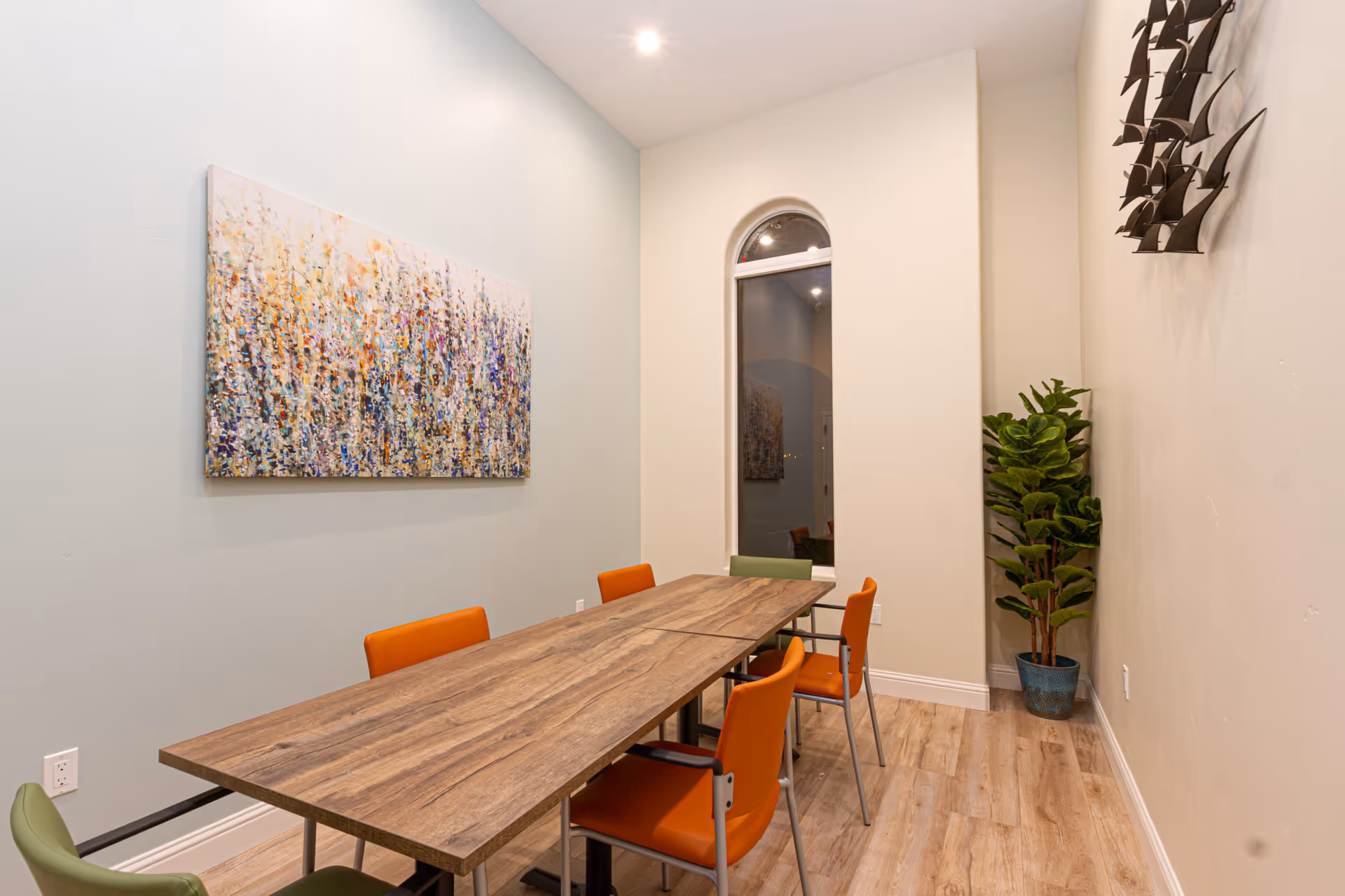 A small meeting or dining room with a long wooden table surrounded by six chairs, four orange and two green. The room has light-colored walls, a tall arched window, a colorful abstract painting on one wall, a metal wall sculpture on another, and a potted plant in the corner. The floor is light wood, and the ceiling has recessed lighting.