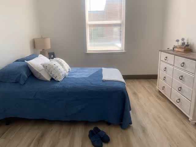 A bedroom with a bed covered in a blue bedspread and several pillows, a nightstand with a lamp and picture frame, a window with blinds, a dresser with decorative items on top, and a pair of slippers on the wooden floor.