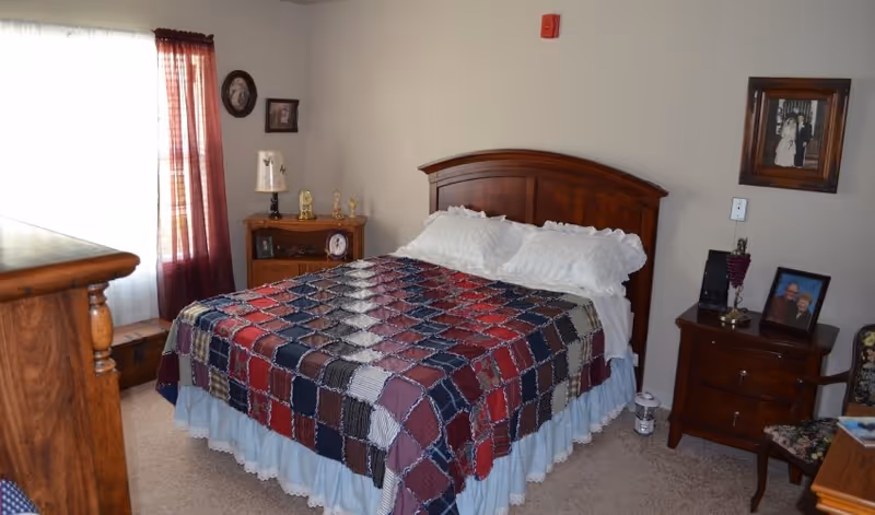 A cozy bedroom with a wooden bed frame and a colorful patchwork quilt on the bed. There are white pillows and a light blue bed skirt. The room has beige walls with framed pictures hanging, a wooden nightstand with a lamp and decorative items, and a window with sheer white curtains and dark red drapes. A wooden dresser and a floral upholstered chair are also visible.