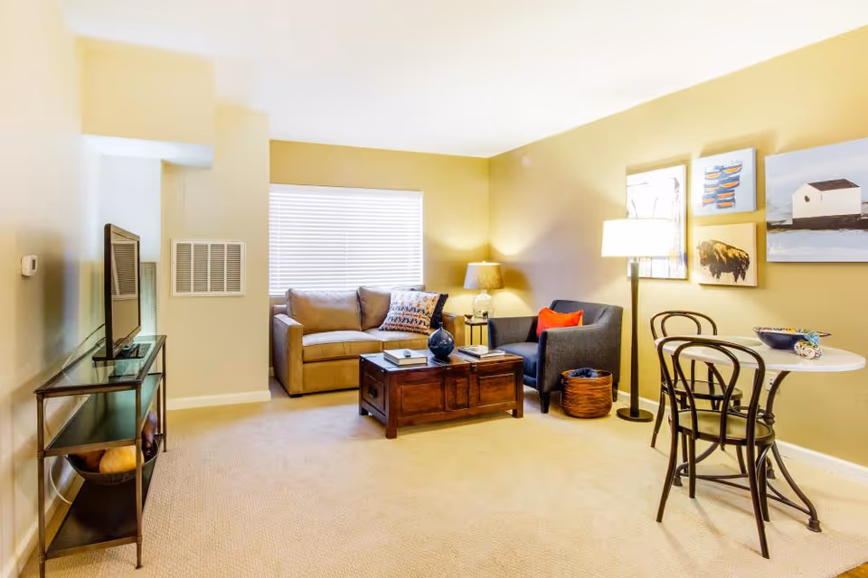 A cozy living room with a beige sofa, a dark armchair with a red pillow, a wooden coffee table, a TV on a glass stand, a small round dining table with two chairs, and wall art. The room is warmly lit with a floor lamp and a table lamp, and has a window with blinds.
