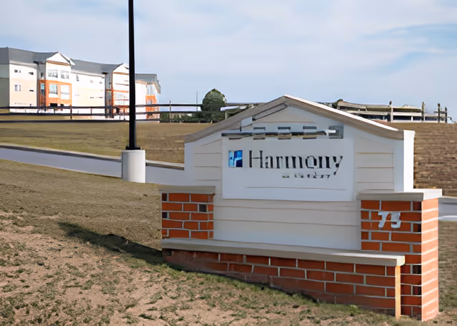 Outdoor view of a sign for Harmony at Hershey facility, with a multi-story building in the background and a clear sky above.