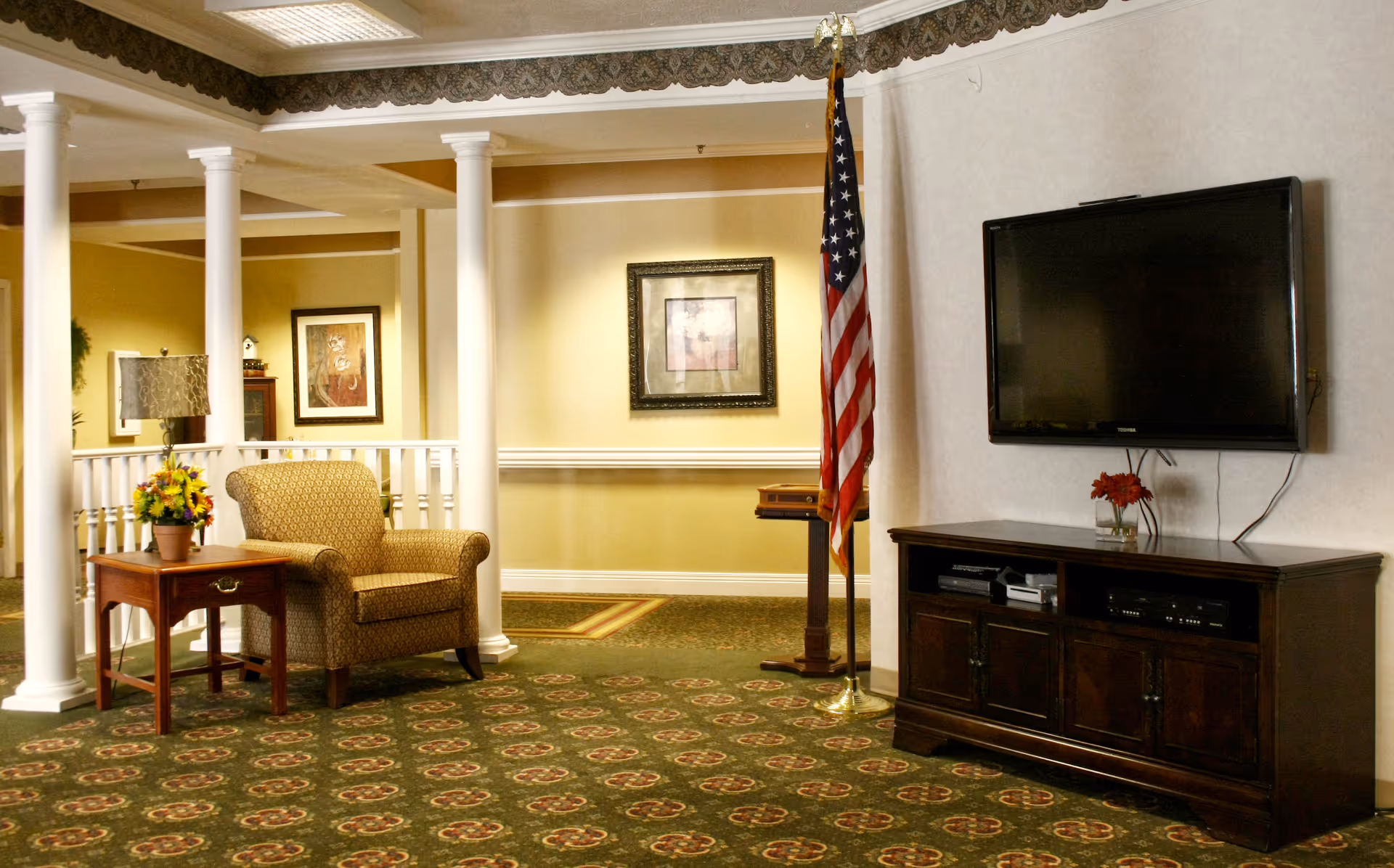 A cozy senior living common area with a patterned armchair next to a wooden side table holding a flower pot and lamp. The room features white columns, framed artwork on the walls, an American flag on a stand, and a flat-screen TV mounted above a dark wooden cabinet. The carpet has a green and red floral pattern.