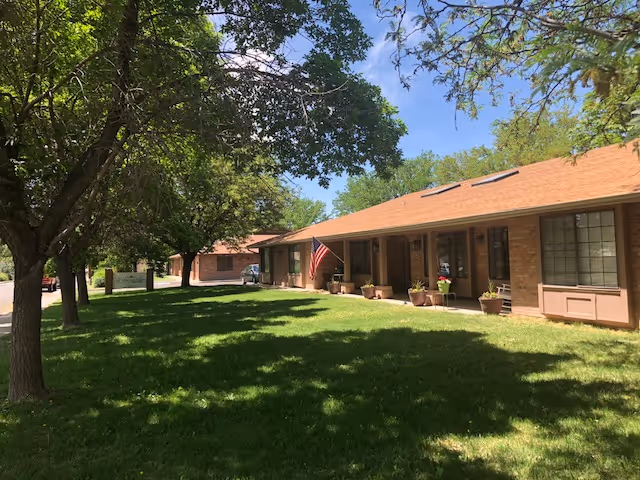 Single-story brick building with a sloped roof and several windows, surrounded by green grass and large trees under a clear blue sky. An American flag is displayed near the entrance.