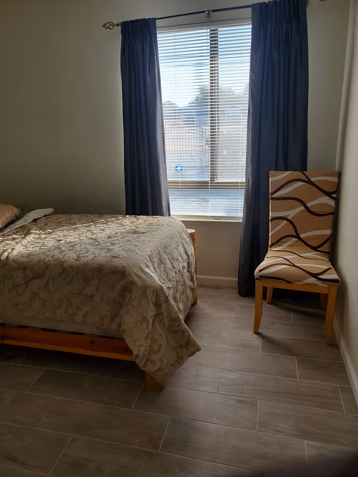 A bedroom with a bed covered in a beige patterned bedspread positioned next to a window with closed blinds and dark curtains. A wooden chair with a patterned cushion is placed against the wall to the right of the window. The floor is tiled with light-colored tiles.