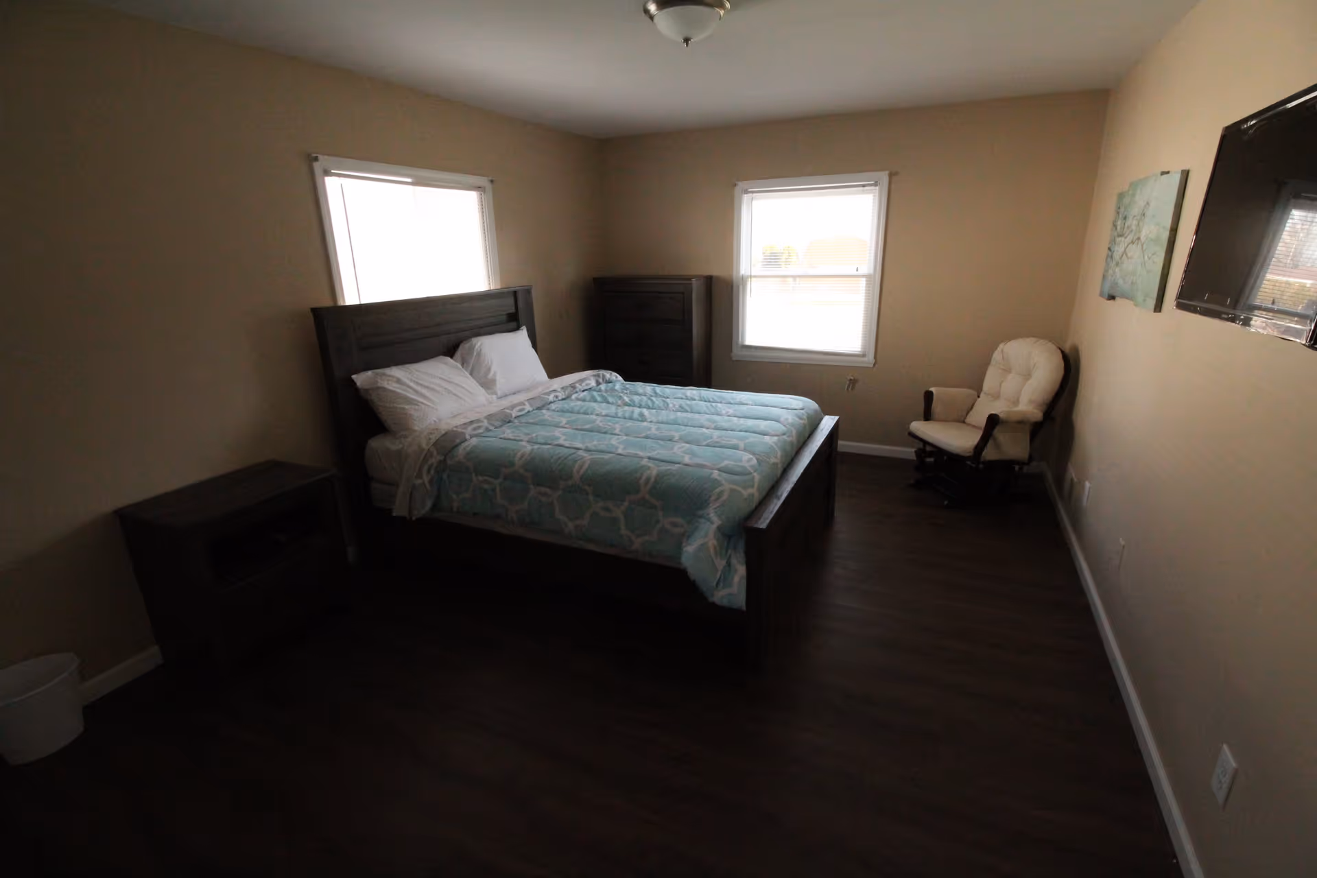 A simple bedroom with a bed made with a patterned comforter, nightstand and dresser, two windows, a wall-mounted TV and a cushioned rocking chair.