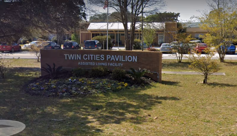 Exterior view of Twin Cities Pavilion assisted living facility showing the building, parking lot with several cars, trees, and a brick sign with the facility name in front.