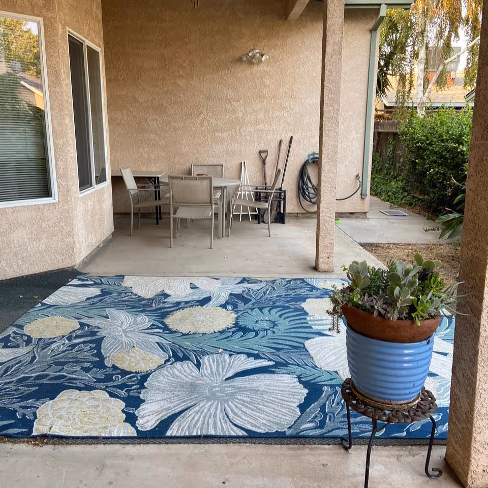 Covered patio with a blue floral outdoor rug, a potted succulent on a small stand, and a table and chairs against a stucco wall.
