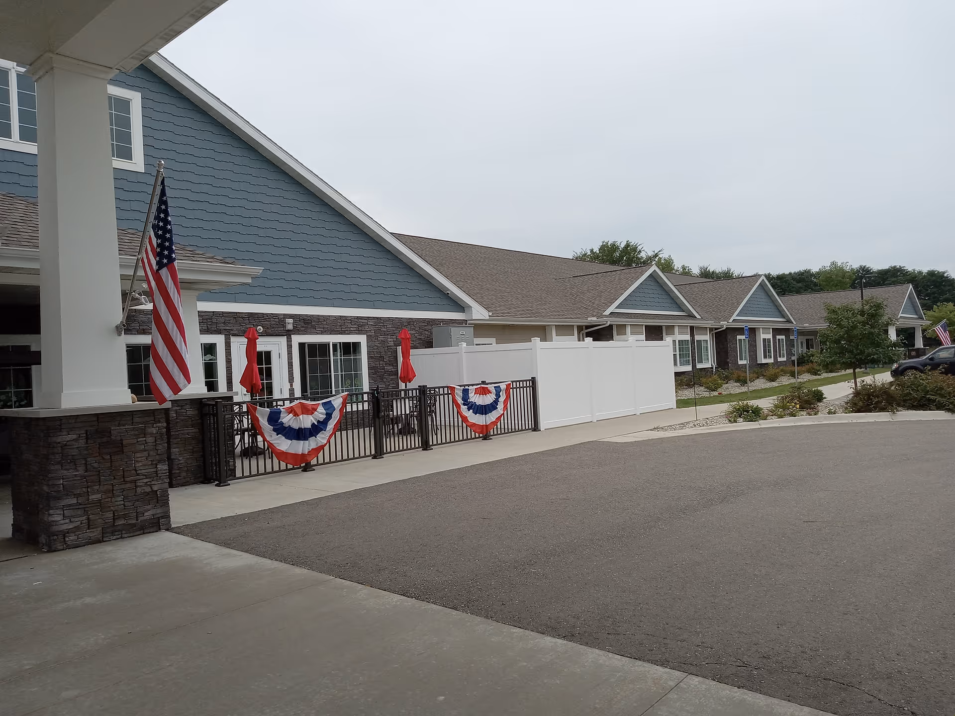 Exterior view of Hampton Manor Hamburg Assisted Living & Memory Care building with blue siding and stone accents. There are American flags and red, white, and blue bunting decorations on the black metal fence. The area includes a paved driveway and some landscaping with bushes and trees.