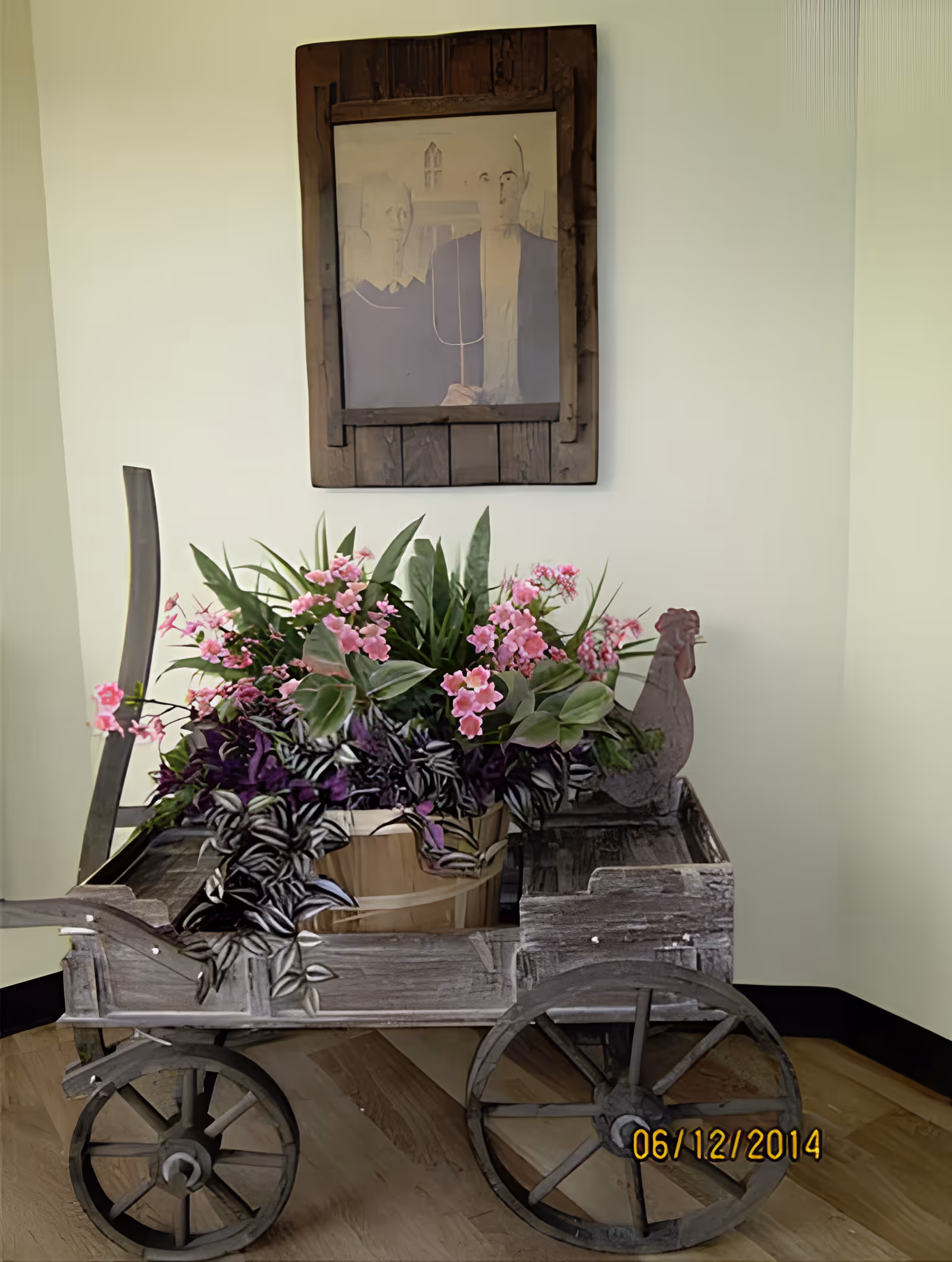 A rustic wooden cart filled with a variety of green and purple plants and pink flowers is placed indoors against a light-colored wall. Above the cart hangs a framed artwork depicting two abstract human figures. The floor is wooden, and the image has a date stamp of 06/12/2014 in the bottom right corner.