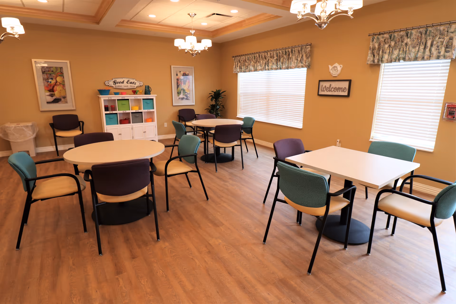 A dining room with three tables and multiple chairs arranged around them. The room has wooden flooring, beige walls, two windows with blinds and floral valances, and ceiling lights. There is a white cabinet with colorful storage bins and decorative signs on the wall, including one that says 'Good Eats' and another that says 'Welcome'.